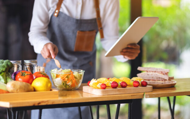 A woman following recipe on digital tablet while cooking salad and sandwich in the kitchen, online learning cooking class concept